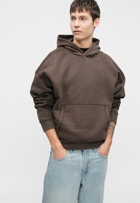 Felpa marrone con cappuccio, tasca frontale, polsini a coste e vestibilità comoda, abbinata a jeans di denim azzurro chiaro.