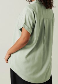 Hellgrüne Bluse mit kurzen Ärmeln und lässigem Schnitt, ausgestattet mit einer weichen Textur, abgerundetem Saum und Faltendetail auf der Rückseite.