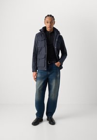 Jachetă parka bleumarin cu multiple buzunare frontale, pulover negru cu fermoar, blugi decolorati, și pantofi negri. Fundal simplu.