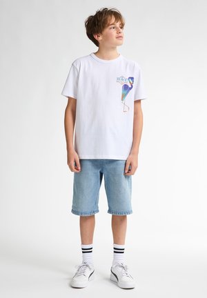 Weißes Baumwoll-T-Shirt mit buntem Vogel-Graphic auf der Brust; kombiniert mit hellblauen Jeansshorts; weiße Sneaker mit schwarzen Akzenten.