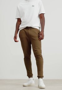 Bruine katoenen joggers met een trekkoord, gecombineerd met een wit shirt met korte mouwen en een klein logo, en witte sneakers.
