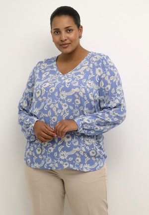 Light blue blouse with floral pattern, V-neck, long sleeves with gathered cuffs, and soft texture. Paired with beige pants.