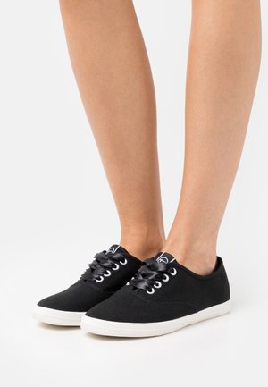 Ben iført sorte lave lærreds sneakers med skinnende sorte snørebånd og hvide gummiydersåler på en ensfarvet hvid baggrund.