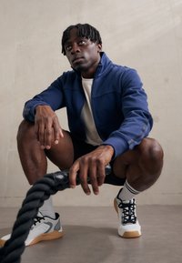Marineblå zip-up jakke, hvid t-shirt, sorte shorts og hvide træningssko med gummisål. Model squatter med et tykt sort reb.