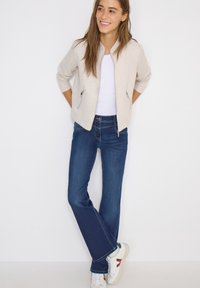 Femme souriante debout contre un mur blanc, portant une veste beige, une chemise blanche, un jean bleu et des baskets blanches avec des accents rouges.