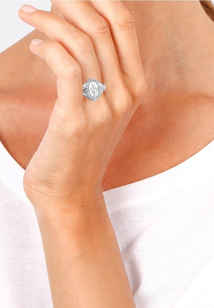 Main portant une bague chevalière religieuse en argent avec une figure gravée, positionnée près du cou sur un haut blanc.