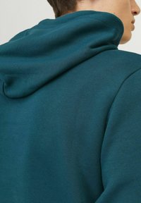 Sudadera de un verde azulado oscuro con una textura suave y capucha ajustable con cordón. Presenta un diseño aerodinámico y acentos acanalados en los bordes.