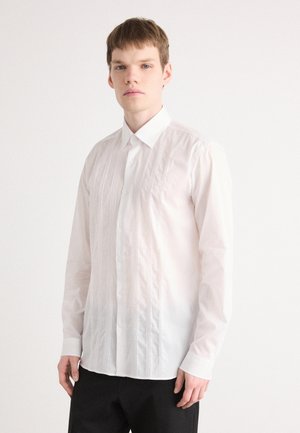 Camisa blanca de manga larga con pliegues verticales, cuello abotonado y una textura suave. Combinada con pantalones oscuros para crear contraste.