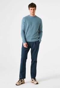 Hellblauer Strickpullover mit gerippten Bündchen und Saum, kombiniert mit dunkelblauen Hosen und beigen Turnschuhen, die eine geschichtete Textur zeigen.