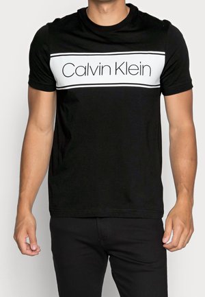 Muž nosící černé krátké tričko s bílým obdélníkovým panelem s nápisem "Calvin Klein" přes hrudník.