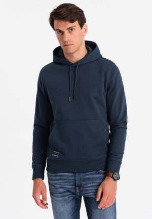 Haut à capuche bleu marine en tissu doux avec une poche avant, des cordons de serrage ajustables et une petite étiquette logo à l'ourlet ; associé à un jean en denim.