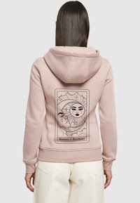 Ljusrosa hoodie med mjuk textur, med en grafisk design av en sol och måne på baksidan, inramad av dekorativa element.