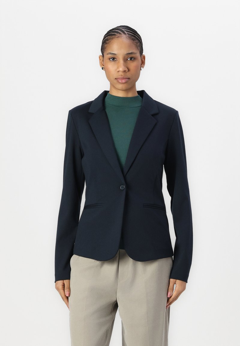 Blazer azul marino con solapas muecas, cierre de un solo botón y dos bolsillos frontales, llevado sobre un cuello de tortuga verde de canalé y pantalones beige.