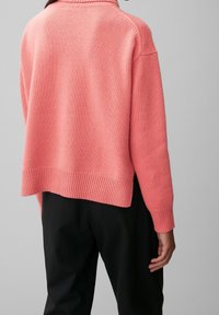 Roze gebreide trui met geribbelde zoom en manchetten, relaxte pasvorm en ronde halslijn, gecombineerd met zwarte broek, gezien vanuit de achterkant.