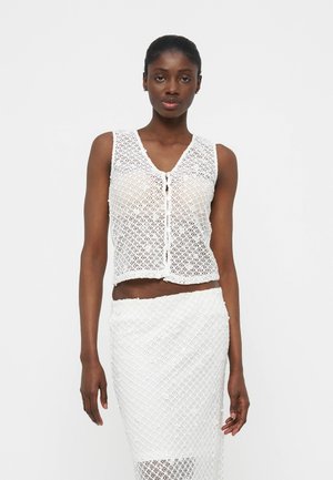 Femme portant un haut en crochet blanc sans manches avec des boutons et une jupe en crochet assortie, debout devant un fond clair uni.