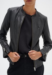 Person iført en sort læderjakke med sølvlynlåse over en sort skjorte, hænder der justerer jakken ved taljen.