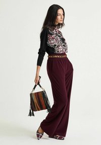 Maglione nero con motivo floreale e maniche lunghe, pantaloni bordeaux a gamba larga, borsa a tracolla con fantasia e nappe, e ballerine multicolori con punta appuntita.