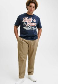 Granatowa bawełniana koszulka z logo "Jack & Jones", zestawiona z beżowymi chinosami i białymi trampkami. Model stoi z rękami w kieszeniach na jednolitym tle.