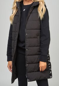 Black puffer vest with a hood, featuring quilted design, front zipper, snap buttons, and side pockets. Worn over a dark sweater.