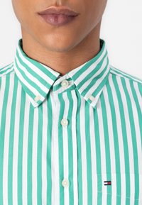 Cămașă cu dungi verzi și albe, cu guler button-down, având nasturi din perlă și un mic logo pe buzunarul de la piept.