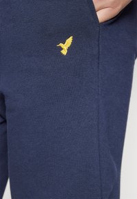 Námořnické jogger kalhoty z měkké tkaniny s přiléhavým střihem, které mají boční kapsy a výrazné žluté vyšívané logo ptáka na horní části stehna.