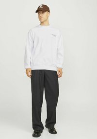 Felpa bianca con maniche lunghe, colletto tondo, dettaglio del logo discreto sul petto, abbinata a pantaloni neri larghi e scarpe nere.