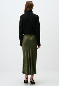 Zwarte gebreide sweater met hoge kraag, gecombineerd met een olijfgroene satijnen rok, die een zachte textuur heeft en een klein splitje aan de achterkant.