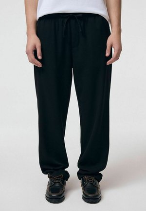 Osoba nosząca luźne czarne spodnie ze sznurkiem i czarne skórzane buty sznurowane, stojąca na jednolitym jasnym tle.