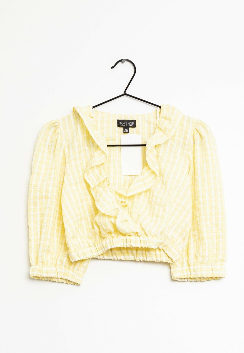 Blusa gialla cropped realizzata in un tessuto leggero con una fantasia a quadri, caratterizzata da un colletto arricciato e polsini elasticizzati. Appesa a un gancio nero.