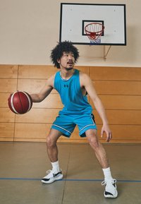 Spiller i en blå ermeløs idrettstopp og matchende shorts, holder en rød og hvit basketball, i et gym med tremurerte vegger og en kurv.