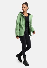 Šviesiai žalia atspari vandeniui striukė su gobtuvu, juodu pamušalu, užtrauktuku užsegamomis kišenėmis, derinama su juodomis džinsinėmis kelnėmis ir juodais auliniais batais.