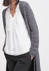 Woman wearing a white blouse with a front tie, black pants, and a long gray textured knit cardigan, with hands in pockets.