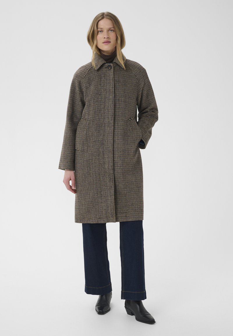 Femme portant un long manteau marron à carreaux, un pantalon foncé et des bottes noires, debout avec une main dans la poche devant un fond blanc uni.