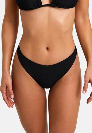 Vrouw draagt zwart geribbeld bikinibroekje en bijpassende top, staat met ontspannen armen langs het lichaam tegen een effen achtergrond.