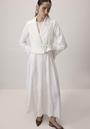 Pikkade tumedate juustega naine kannab valget vöökohaga pintsakut ja pikka seelikut, mis on kokku sobitatud pruunide terava ninaga kinnohubidega, ühtlase taustaga.