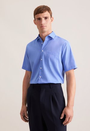 Mann trägt ein hellblaues kurzärmliges Hemd mit Knopfleiste und Brusttasche sowie dunkle, plissierte Hose, steht vor einem schlichten Hintergrund.