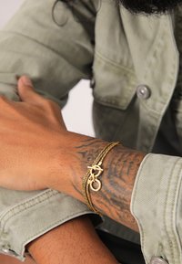 Goldarmband mit verflochtenen Ketten und einem dekorativen Verschluss, getragen an einem tätowierten Handgelenk, im Hintergrund ist der Ärmel einer hellgrünen Jacke zu sehen.