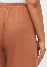 Pantalons larges de couleur marron, légers et texturés, avec une taille élastique, montrant un gros plan du dos et une main gauche reposant sur le tissu.