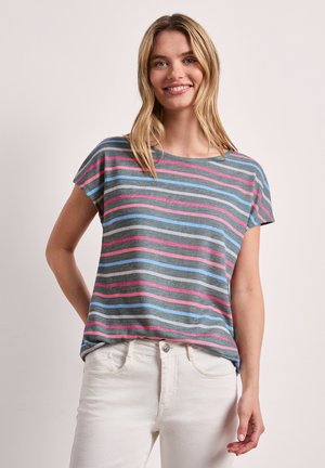 Lachende vrouw met blond haar, die een grijs T-shirt draagt met roze, blauwe en witte horizontale strepen en een witte broek, staande met één hand achter haar rug.