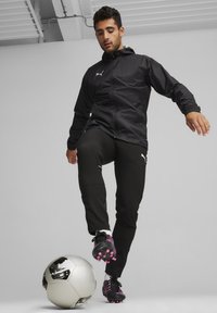 Schwarze Jacke mit Kapuze, Reißverschluss vorne, elastische Bündchen; schwarze Hose mit weißen Akzenten; schwarze Fußballschuhe mit pinken Details; einen Fußball treten.