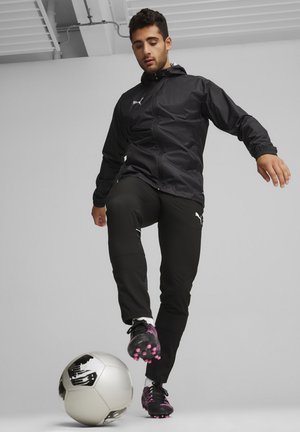 Black jacket with hood, zippered front, elastic cuffs; black pants with white accents; black soccer shoes with pink detailing; kicking a soccer ball.