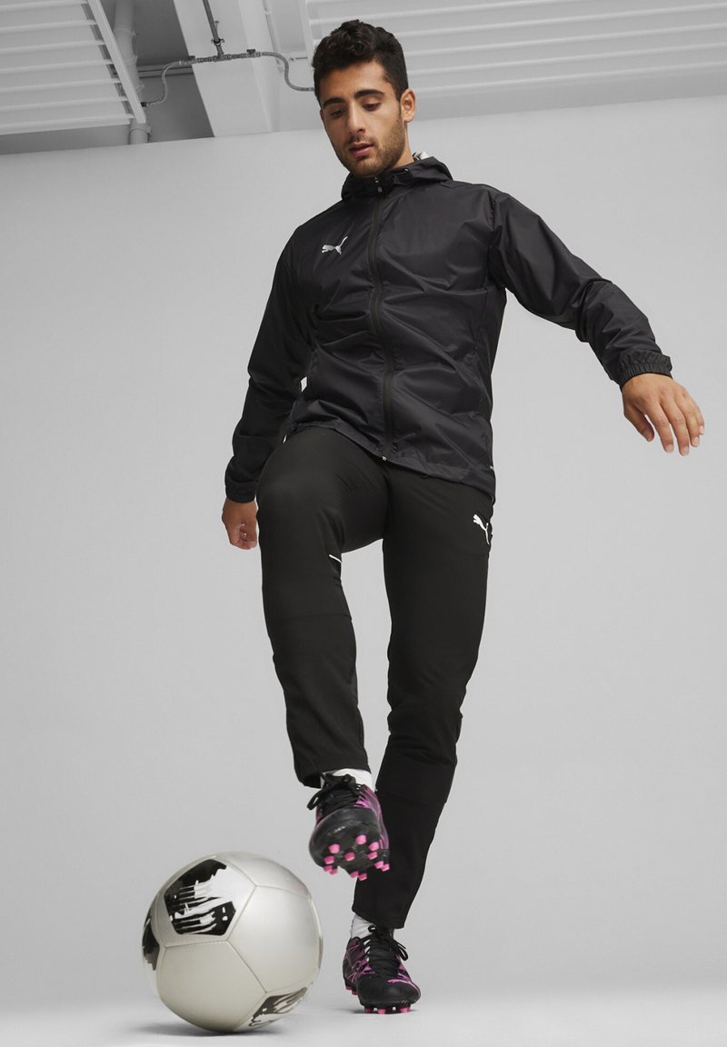 Schwarze Jacke mit Kapuze, Reißverschluss vorne, elastische Bündchen; schwarze Hose mit weißen Akzenten; schwarze Fußballschuhe mit pinken Details; einen Fußball treten.