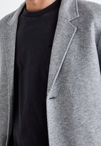 Uomo che indossa un blazer grigio testurizzato sopra una maglietta nera a girocollo, visibile dal collo fino a metà torace.