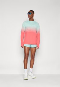Langearm sportshirt in een gradient van mint naar koraal, gecombineerd met mintkleurige shorts en witte sneakers. Voorzien van ademend materiaal en een atletische pasvorm.