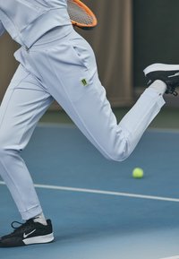 Gaiši zils sporta tērps ar rāvējslēdzējam kabatām un melnām akcentiem, kombinēts ar melniem sporta apaviem. Tenisa bumba kustībā uz zila laukuma.