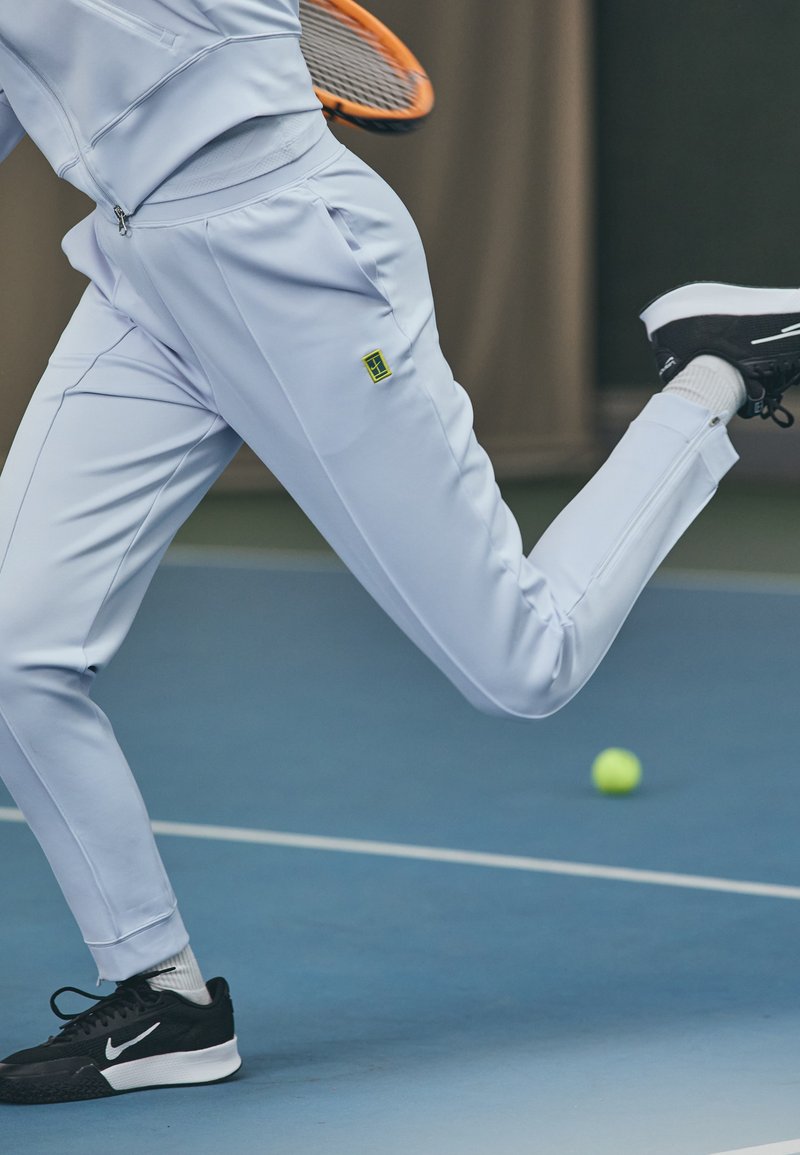 Gaiši zils sporta tērps ar rāvējslēdzējam kabatām un melnām akcentiem, kombinēts ar melniem sporta apaviem. Tenisa bumba kustībā uz zila laukuma.
