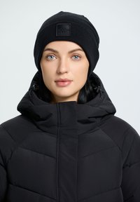 Chaqueta acolchada negra con cuello alto y acabado mate, que presenta paneles cosidos. Gorro negro con diseño ribeteado texturizado y parche con logo.
