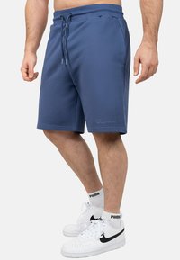 Shorts sportivi blu navy con vita regolabile tramite cordino, tasche laterali e logo ricamato sull'orlo. Indossati con sneakers bianche e calze.