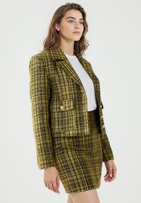 Geel en zwart tweed blazer met een gestructureerd ontwerp, voorzien van een uitgesneden kraag, gouden knopen en twee voorkeurszakken. Gepaard met een bijpassende rok.