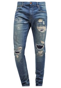 Vaqueros azules de mezclilla con un corte ajustado, detalles desgastados con parches rasgados y tela oscura contrastante visible debajo.